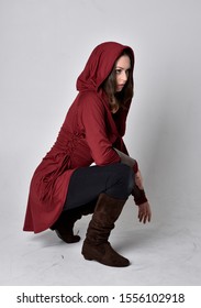 full length portrait of a brunette girl wearing a red fantasy tunic with hood. Seated pose on a white studio background.