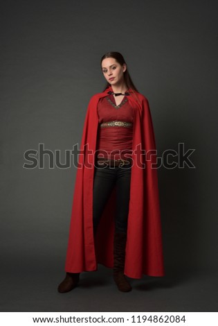 full length portrait of brunette girl wearing red medieval costume and cloak. standing pose   on grey studio background.