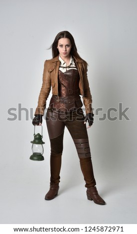 
full length portrait of brunette  girl wearing brown leather steampunk outfit. standing pose holding a gas lantern on grey studio background.