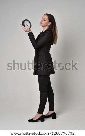 
full length portrait of a brunette girl wearing long black coat, standing pose holding a small clock on grey studio background.