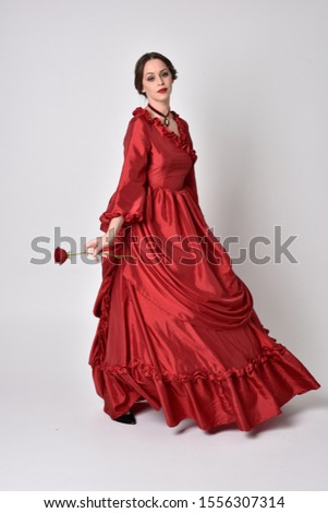 full length portrait of a brunette girl wearing a red silk victorian gown. Standing pose, holding a rose,  on a white studio background.