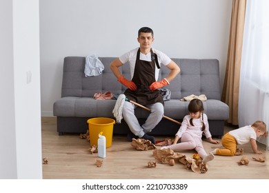 Full Length Portrait Of Brunette Man Wearing Brown Apron Holding Mop And Sitting On Sofa, Doing Domestic Chores, Cleaning Apartment, Don't Know Where To Start Cleaning Up The Mess.