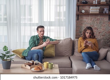 Full Length Portrait Of Busy Married Couple Using Smartphones With Concentration. They Are Sitting On Sofa At Home