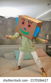 Full Length Portrait Of Carefree Black Child Playing With Cardboard Box And Jumping On Bed At Home