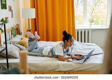 Full length portrait of carefree teenage girl using laptop on bed in cozy room, copy space