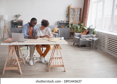 Full length portrait of caring African-American father helping son doing homework or studying while sitting together at desk in home interior, copy space