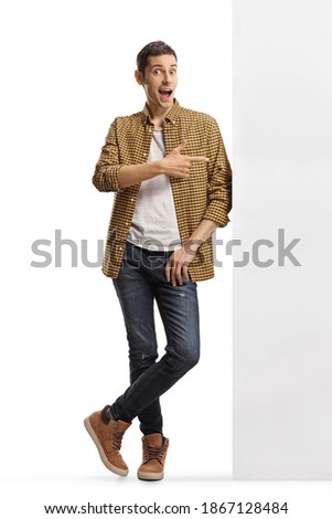 Full length portrait of a casual man in checkered shirt and jeans leaning on a wall and pointing to the side isolated on white background
