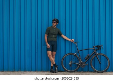 Full length portrait of caucasian male athlete in sport outfit keeping hand on black bike while leaning on blue wall. Cyclist standing outdoors and looking aside. - Powered by Shutterstock