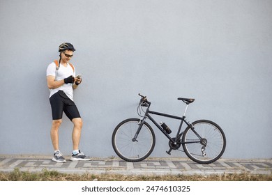 Full length portrait of Caucasian male athlete in sport outfit near black bike while leaning on wall. Cyclist standing outdoors and looking at smartphone. - Powered by Shutterstock