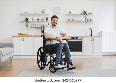 Full length portrait of Caucasian wheelchair user smiling at camera while resting in open-plan kitchen of modern apartment. Cheerful young male in casual clothes spending pastime indoors. - Powered by Shutterstock