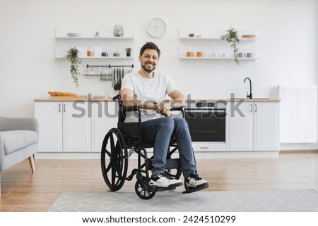 Full length portrait of Caucasian wheelchair user smiling at camera while resting in open-plan kitchen of modern apartment. Cheerful young male in casual clothes spending pastime indoors.