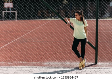 Full length portrait of charming young lady with dark hair standing near chain link fence at modern stadium. Relaxed woman in sport outfit spending free time at sport ground. - Powered by Shutterstock