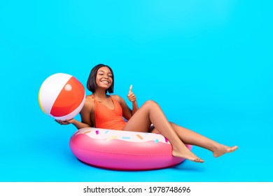 Full length portrait of cheerful black lady in swimwear sitting in inflatable ring, holding beach ball and showing thumb up gesture, recommending summer vacation on blue studio background, copy space