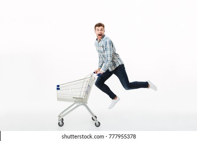 Full length portrait of a cheerful man jumping with a shopping trolley isolated over white background