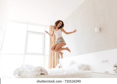 Full length portrait of a cheerful woman in earphones singing and jumping on bed indoors