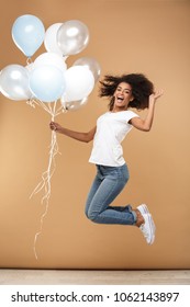 Full length portrait of a cheerful young african woman jumping while holding bunch of air balloons isolated over beige background