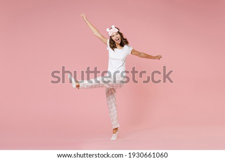 Full length portrait of cheerful young woman in white pajamas home wear sleep mask standing on toes rising spreading hands leg rest at home isolated on pink background studio. Relax good mood concept