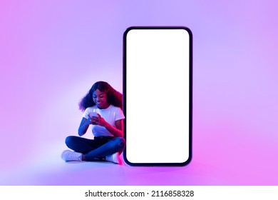 Full Length Portrait Of Cheery Black Woman Sitting Near Giant Smartphone With Mockup For Your App Design, Using Mobile Device In Neon Light. Cellphone Touchscreen Template For Website Or Ad