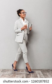 Full Length Portrait Of A Confident African Business Woman Wearing Suit Standing Over Gray Background, Holding Takeaway Coffee, Carrying Laptop