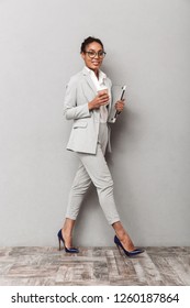 Full Length Portrait Of A Confident African Business Woman Wearing Suit Standing Over Gray Background, Holding Takeaway Coffee, Carrying Laptop