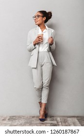 Full Length Portrait Of A Confident African Business Woman Wearing Suit Standing Over Gray Background, Holding Takeaway Coffee, Carrying Laptop