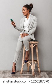 Full Length Portrait Of A Confident African Business Woman Wearing Suit Sitting On A Chair Over Gray Background, Holding Takeaway Coffee, Using Mobile Phone