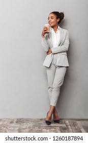 Full Length Portrait Of A Confident African Business Woman Wearing Suit Standing Over Gray Background, Holding Cup Of Takeaway Coffee