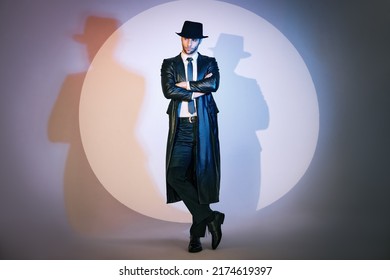 Full length portrait of confident handsome man in black coat and hat with crossed arms posing on studio in the spotlight. Noir film style. Private detective, spy, investigation concept - Shutterstock ID 2174619397