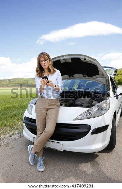Full length portrait of confident middle aged woman\
using her mobile phone while standing on the road at her broken\
down car.