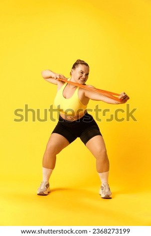 Full length portrait of confident plus size sports woman in tracksuit training hard with sport elastic bands for hands over yellow background. Concept of sport, hobby, health, lifestyle, workout