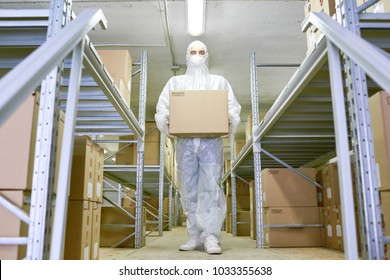 Full Length Portrait Of Confident Worker Wearing Safety Mask And Coverall Carrying Cardboard Box And Walking Along Shelves Of Pharmaceutical Factory Warehouse