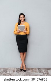 Full Length Portrait Of A Confident Young Business Woman Holding Digital Tablet While Standing Against Gray Background