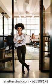 Full Length Portrait Of Confident Young Woman Standing In Doorway Of Office With Her Arms Crossed. Creative Female Executive At Startup With People Working In Background.