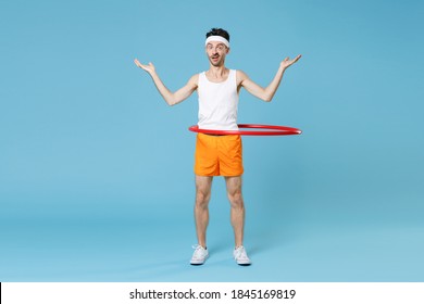 Full Length Portrait Confused Young Man With Skinny Body Sportsman In Headband Shirt Shorts Training With Hula Hoop Spreading Hands Isolated On Blue Background. Workout Gym Sport Motivation Concept