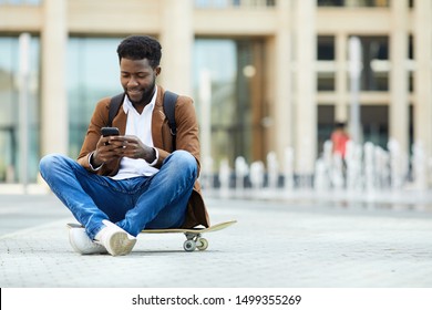 Full length portrait of contemporary African-American man using smartphone and smiling while sitting cross legged on skateboard outdoors, copy space - Powered by Shutterstock