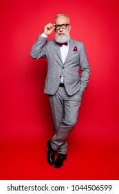 Full Length Portrait Of Cool Stunning Stylish Trendy Old Man In Checkered Jacket And Pants, Holding Hand In Pocket And Eyelet Of Glasses On His Face, Standing Leg By Foot Over Red Background