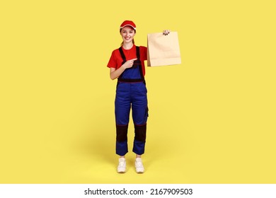 Full Length Portrait Of Courier Woman Giving Ordered Parcel, Delivering Food In Paper Bag, Post Mail Services, Wearing Overalls And Red Cap. Indoor Studio Shot Isolated On Yellow Background.