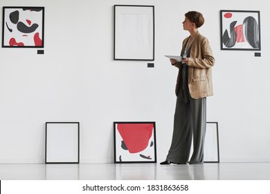 Full Length Portrait Of Creative Elegant Woman Looking At Paintings Hanging On White Wall While Visiting Modern Art Gallery Exhibition, Copy Space