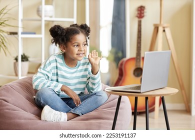 Full length portrait of cute black girl waving to laptop camera and smiling while enjoying online lesson from home sitting cross legged in comfy bean bag chair, copy space