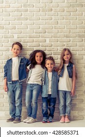 Full length portrait of cute little kids in stylish jeans clothes hugging, looking at camera and smiling, standing against white brick wall