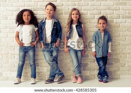 Full length portrait of cute little kids in stylish jeans clothes looking at camera and smiling, standing against white brick wall