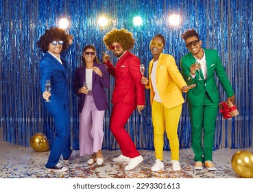 Full length portrait of dancing men and women drinking alcohol in sunglasses and colourful suits in gangnam style on blue shiny background. Friends in funny curly wigs at night club disco party.