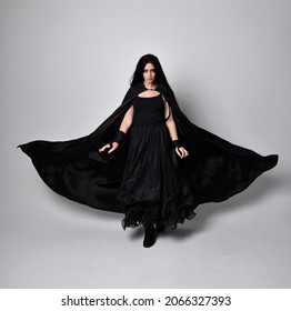 Full length portrait of dark haired girl wearing a witch black flowing gown and  fantasy cloak.   Standing pose with gestural movements, isolated on studio background. - Powered by Shutterstock