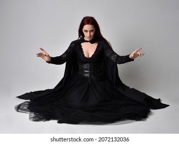 Full length portrait of dark haired girl wearing a fantasy witch black costume,  sitting pose with   gestural movements, isolated on studio background.