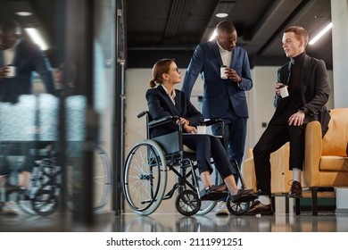 Full length portrait of diverse business team with young woman in wheelchair chatting to male colleagues in modern office