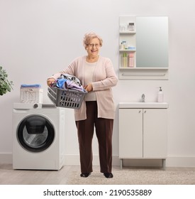 Full Length Portrait Of An Elderly Woman Holding A Laundry Basket With Clothes Inside A Bathroom