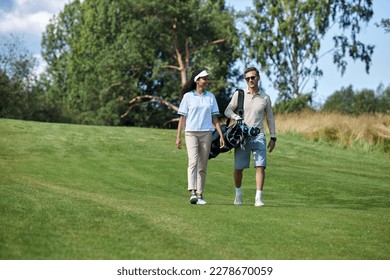 Full length portrait of elegant sporty couple carrying golf bag walking on green field towards camera and chatting, copy space - Powered by Shutterstock