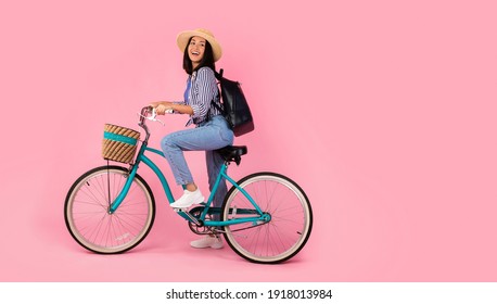 Full length portrait of excited asian woman wearing summer hat and black backpack riding vintage bicycle with wicker basket looking back at free copy space isolated on pink studio background