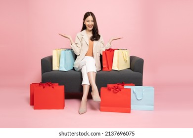 Full Length Portrait Of Excited Asian Woman Sitting On Sofa With Shopping Bags Isolated On Pink Background, Shopper Or Shopaholic Concept