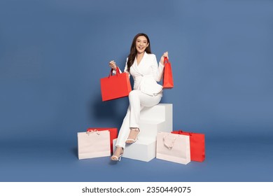 Full length portrait of Excited Asian businesswoman sitting on white stair with shopping bags isolated on blue background, Shopper or shopaholic concept - Powered by Shutterstock